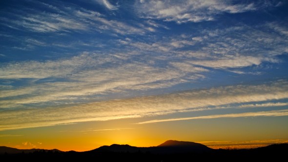 Sunset sky with mountains silhouetted in distance and high, white clouds against blue and yellow sky