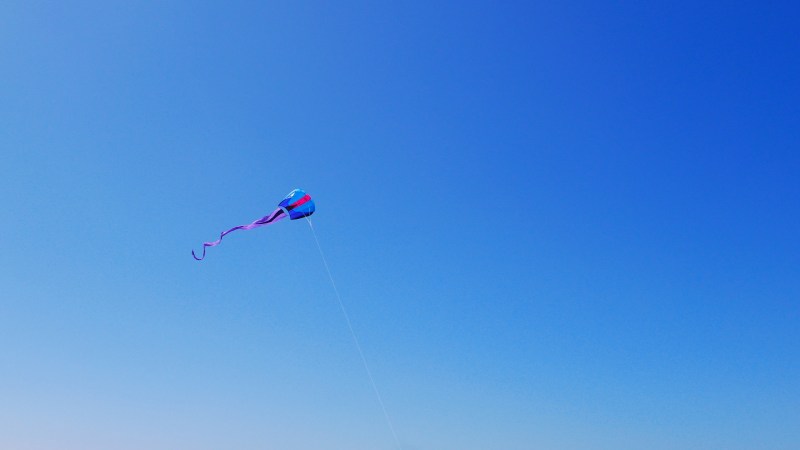 Blue and red kite with purple tail soaring in blue sky
